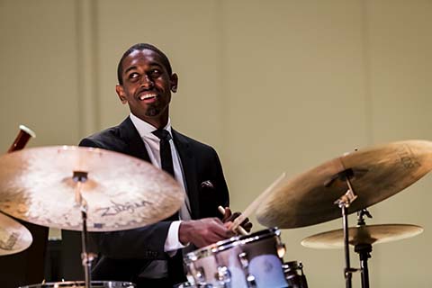 Man in a dark suit smiles while playing the drums during a performance