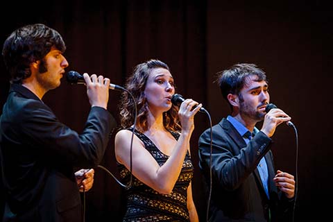 Three vocalists perform with microphones in their hands on stage