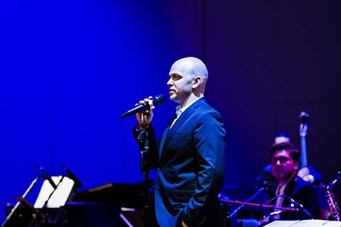 Man in a dark colored suit holds a microphone while musicians look on in the background